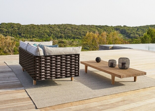 bruine loungeset met grijze kussens en bruine houten tafel schuin achterzijde op een tapijt op een platform met berg landschap